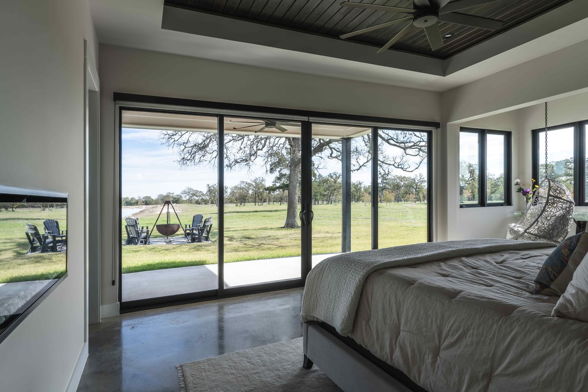 Bedroom view with a bed facing large sliding glass doors opening to a scenic outdoor area with chairs around a fire pit | J Bryant Boyd