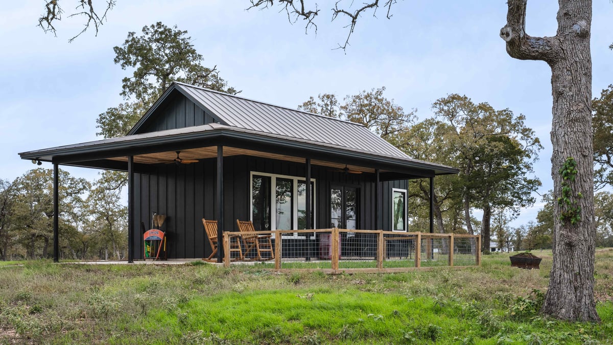 Exterior view of a small black cabin with a metal roof, a wooden porch, and surrounding trees | J Bryant Boyd