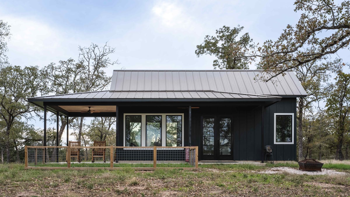 Front view of a small black cabin with a metal roof, wooden porch, and large windows, set in a natural landscape | J Bryant Boyd