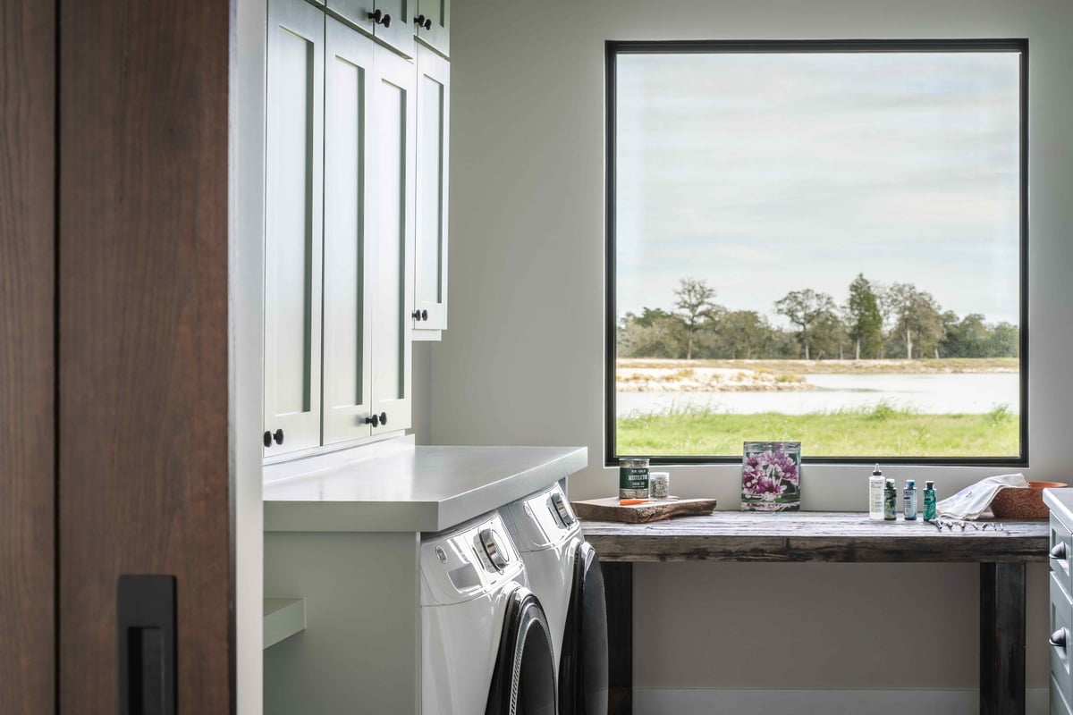 Laundry room with white cabinets, a washer and dryer, and a large window overlooking a serene outdoor landscape | J Bryant Boyd