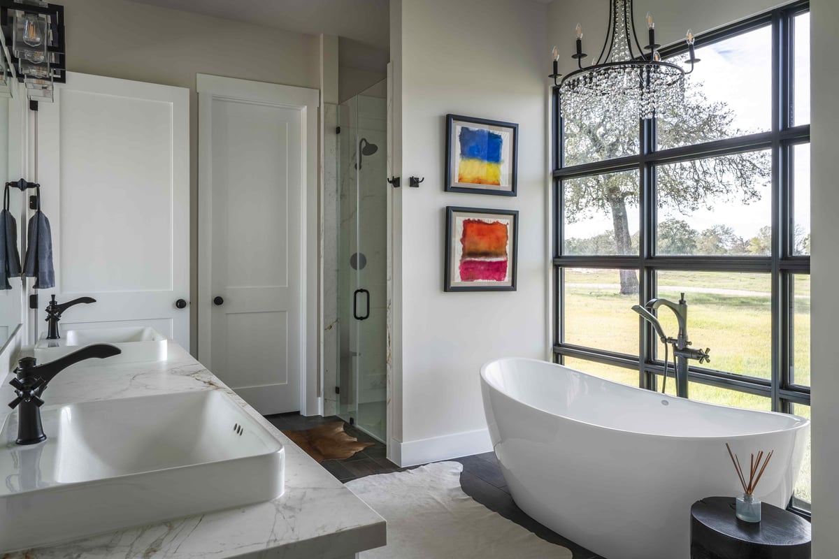 Luxurious bathroom with dual sinks, a freestanding tub by large windows, and a chandelier, featuring colorful abstract paintings | J Bryant Boyd