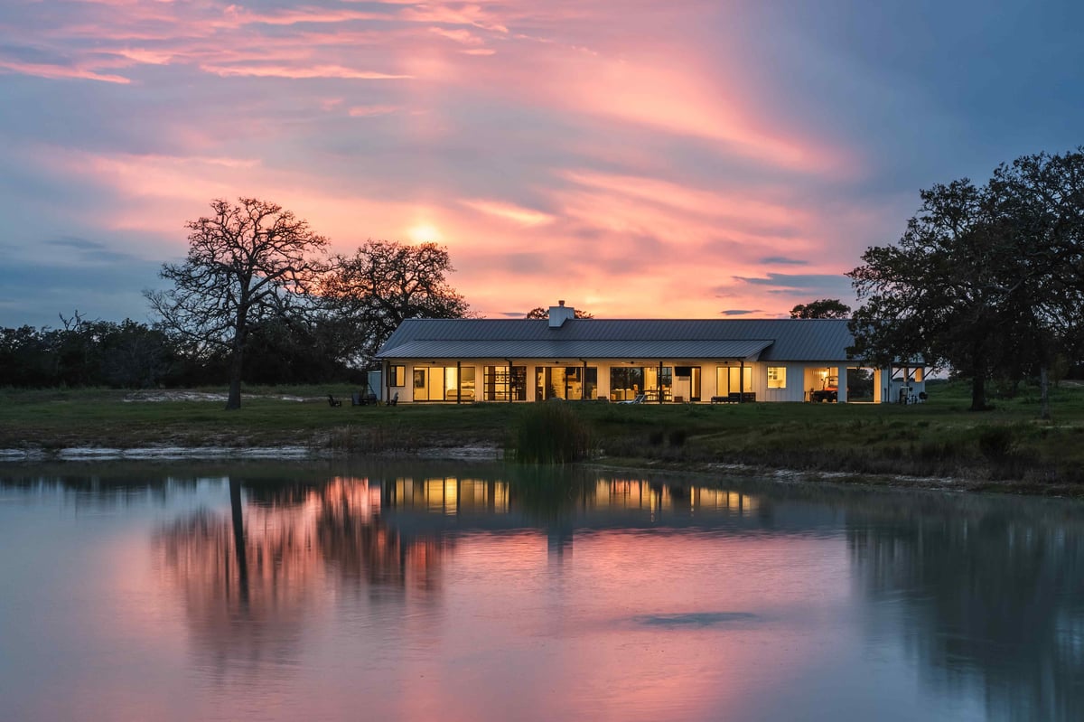 Modern farmhouse exterior at sunset, reflecting on a nearby pond. | J Bryant Boyd